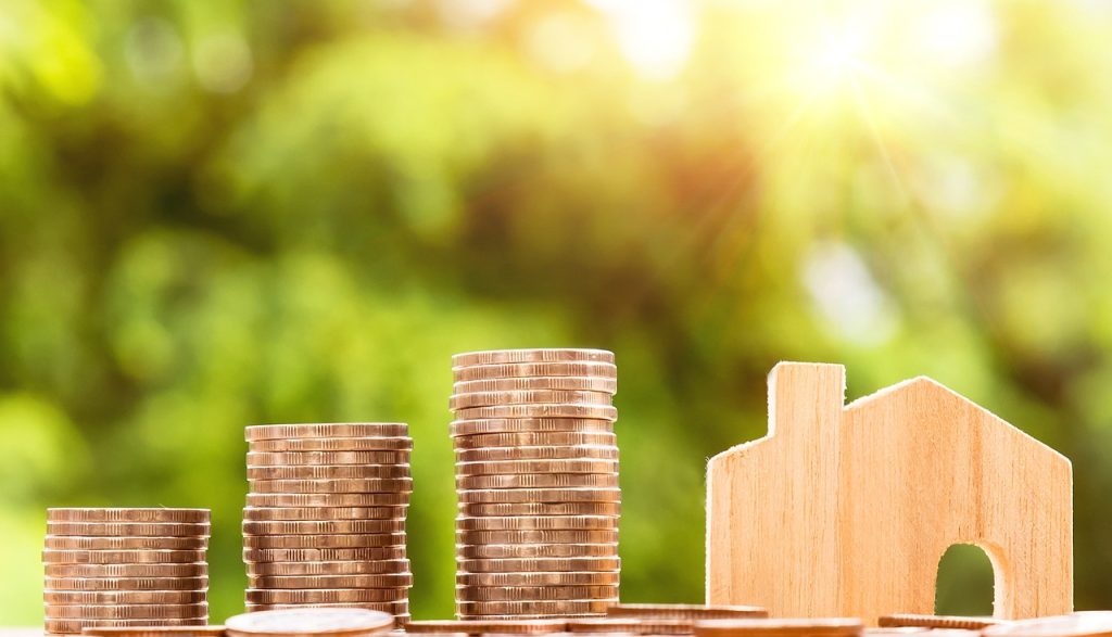 Real Estate Investment Strategy visial aid using stacked coins next to a wooden cut out of a house. the background is a blurred sunbeam peaking through the trees