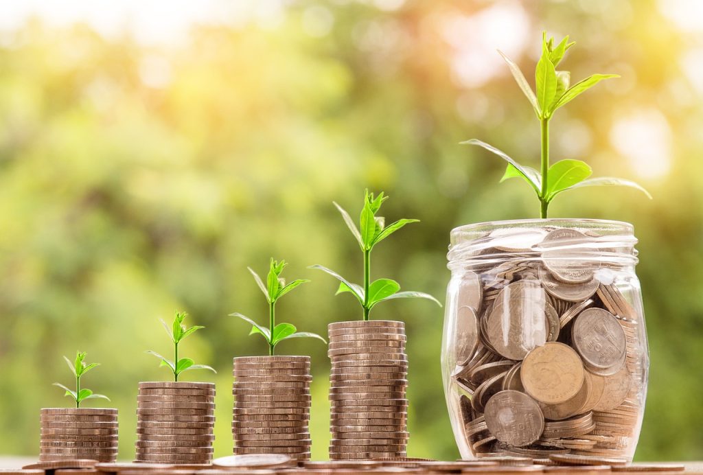a stack of coins gets larger from left to right until the stack is a jar of coins. There are "sprouts" of a generic leafy green growing with each stack. The background is a blur of greenery, like bushes, used as a visual aid for the secret to building wealth.